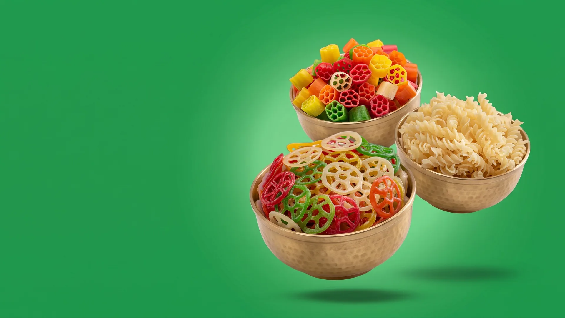 Brass bowls of Suvidha Papad Wheel fryums, Fool fingers and Spring pasta floating on a green backdrop
